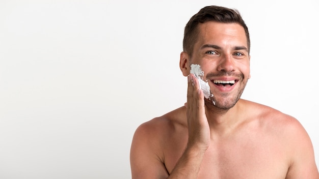 Retrato de joven sin camisa aplicando espuma de afeitar de pie contra la pared blanca