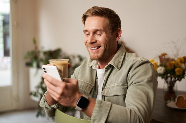 Foto gratuita retrato de un joven carismático sonriente sentado en una cafetería bebiendo capuchino mirando a su