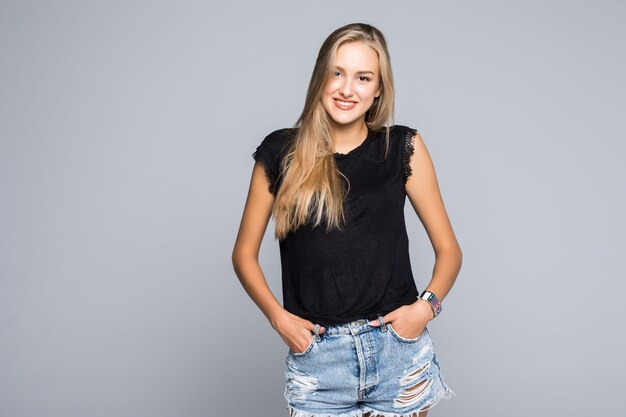 Retrato de una joven casual sosteniendo sus pulgares en los bucles de sus pantalones vaqueros cortos mientras sonríe para la cámara sobre un fondo gris