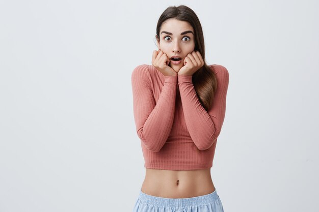 Retrato de joven caucásica guapa con cabello largo oscuro en top rosa de moda y pantalones cortos deportivos azules tomados de la mano cerca de la boca con expresión asustada