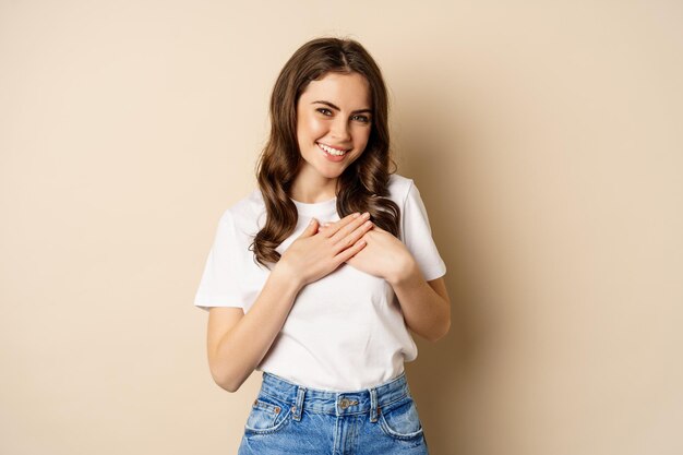 Retrato de una joven caucásica con las manos en el corazón y sonriendo, soñando despierto, pensando en algo conmovedor, posando sobre un fondo beige.