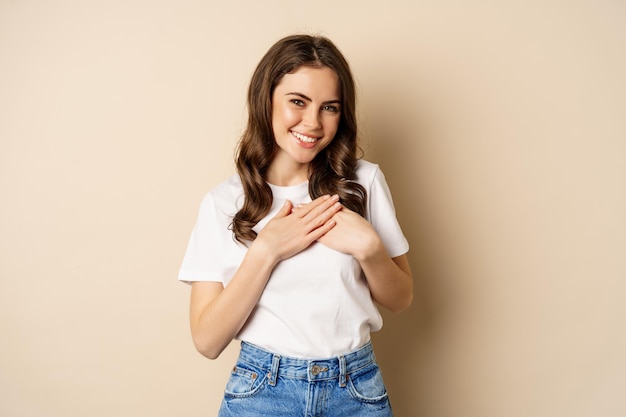 Retrato de una joven caucásica con las manos en el corazón y sonriendo, soñando despierto, pensando en algo conmovedor, posando sobre un fondo beige.