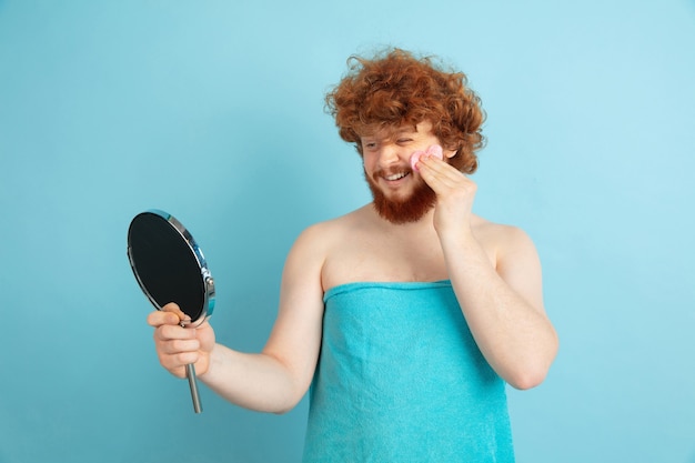 Foto gratuita retrato de joven caucásico en su día de belleza y rutina de cuidado de la piel. modelo masculino con cabello rojo natural aplicando crema hidratante, aceites en la cara. cuidado del cuerpo y la cara, concepto de belleza natural, masculina.