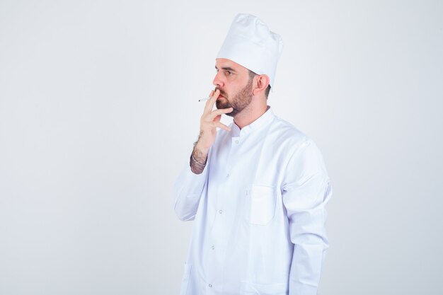 Retrato de joven chef fumando cigarrillos en uniforme blanco y mirando pensativo vista frontal