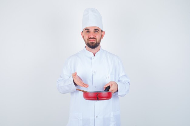 Retrato de joven chef masculino mostrando una sartén vacía con cuchara de madera en uniforme blanco y mirando abatido vista frontal