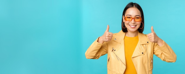 Retrato de una joven china asiática moderna con gafas de sol que usa ropa de abrigo de moda que muestra los pulgares hacia arriba y sonríe complacida recomendando un fondo azul suave