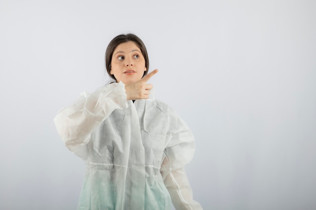 Retrato de joven científica doctora en bata de laboratorio defensiva apuntando hacia arriba.