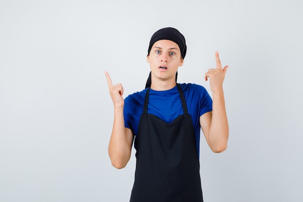 Retrato de joven cocinero adolescente apuntando hacia arriba en camiseta, delantal y mirando desconcertado vista frontal