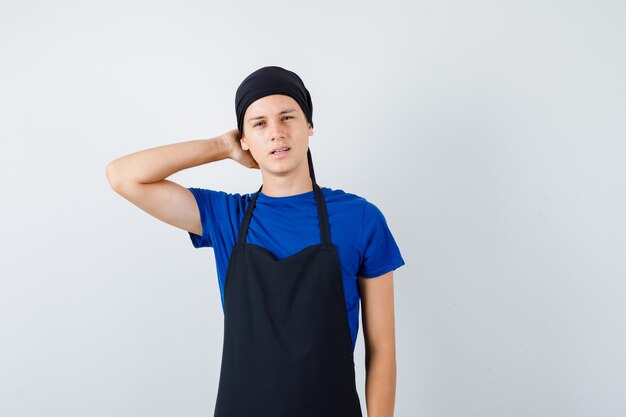 Retrato de joven cocinero adolescente con la mano detrás de la cabeza en camiseta, delantal y mirando pensativo vista frontal