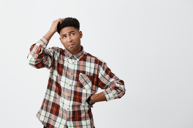 Retrato de joven confuso guapo guapo de piel oscura con peinado afro en camisa a cuadros sosteniendo la cabeza con la mano, mirando a un lado con expresión satisfecha, no sé qué hacer con las deudas.