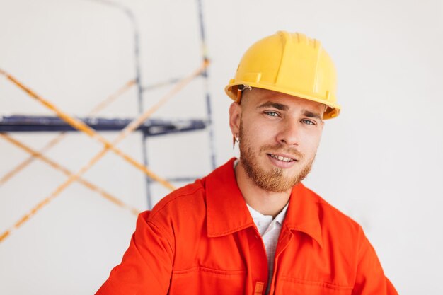 Retrato de joven constructor en ropa de trabajo naranja y casco amarillo con lápiz detrás de la oreja mirando soñadoramente en cámara con andamios en el fondo