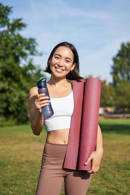 Retrato de una joven coreana delgada y saludable haciendo ejercicio en el parque de pie con una botella de agua y