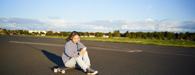 Foto gratuita retrato de una joven coreana sentada en su patineta en la carretera mirando su teléfono inteligente chateando