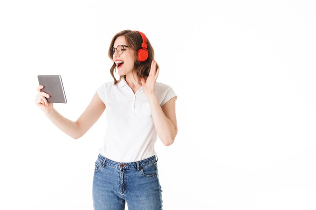 Retrato de joven dama alegre en anteojos y auriculares de pie y felizmente mirando en tableta sobre fondo blanco aislado