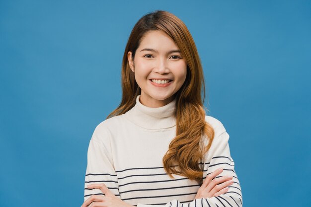 Retrato de una joven dama de Asia con expresión positiva, con los brazos cruzados, una amplia sonrisa, vestida con ropa informal y mirando al frente sobre la pared azul