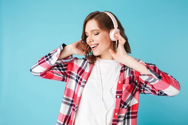 Retrato de joven dama bonita en camisa bailando mientras escucha música en auriculares sobre fondo rosa