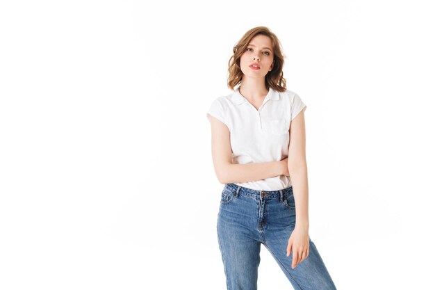 Retrato de joven dama pensativa de pie en camiseta y jeans y mirando cuidadosamente en cámara sobre fondo blanco aislado