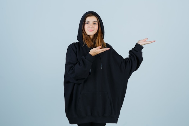 Retrato de joven dando la bienvenida en sudadera con capucha de gran tamaño, pantalones y mirando feliz vista frontal