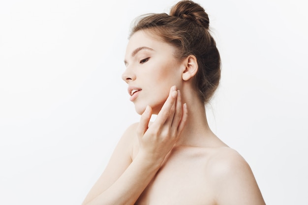 Retrato de una joven y delgada mujer europea delgada con cabello castaño en elegante peinado de moño y hombros desnudos, gira la cabeza a un lado con los ojos cerrados, tocando suavemente la cara con la mano, relajándose y