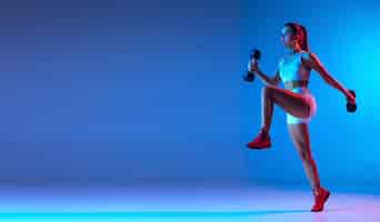 Foto gratuita retrato de una joven deportista entrenando con pesas aisladas sobre fondo azul en neón