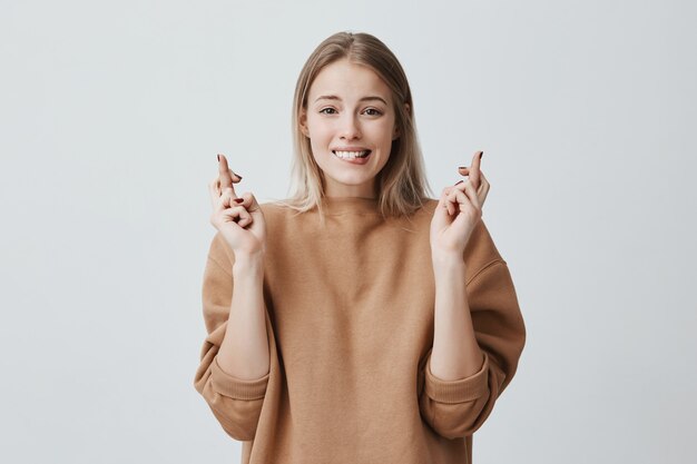 Retrato de una joven deseosa en ropa casual con cabello rubio teñido, cruzando los dedos, mordiéndose el labio inferior, sintiéndose nerviosa ante un evento importante. Lenguaje corporal