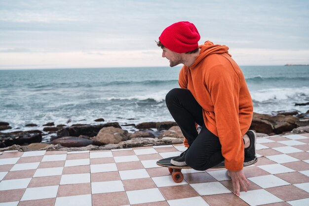 Retrato de joven divirtiéndose con su patineta y practicando sus trucos con el mar en el espacio.