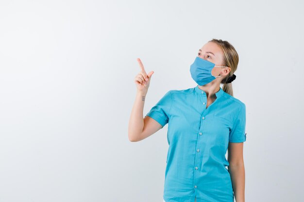 Retrato de joven doctora apuntando hacia arriba en uniforme y mirando pensativo