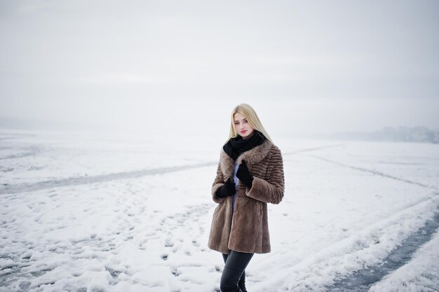 Retrato de joven elegancia chica rubia en un abrigo de piel de fondo río brumoso en el hielo de invierno
