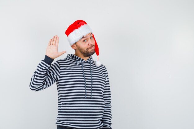 Retrato de joven elegante saludo con la mano abierta en sudadera con capucha, gorro de Papá Noel y mirando alegre