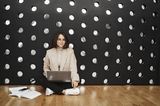 Retrato de una joven elegante sentada en el suelo y en una pared inclinada mientras estudiaba sostener una laptop en el regazo aprendiendo material nuevo para los cursos lectura de libros y hojas de trabajo cámara de sonrisas