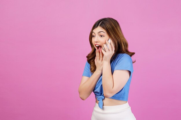 Retrato de una joven emocionada con una camiseta informal hablando por teléfono móvil aislada sobre un fondo rosa