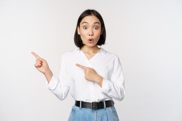 Retrato de una joven y emocionada dama de oficina asiática señalando con el dedo a la izquierda con un descuento que muestra un banner de venta de pie sobre fondo blanco