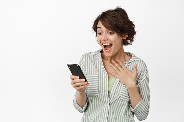 Retrato de una joven emocionada y sorprendida mirando el teléfono móvil feliz, riendo asombrada por el mensaje, de pie contra un fondo blanco.