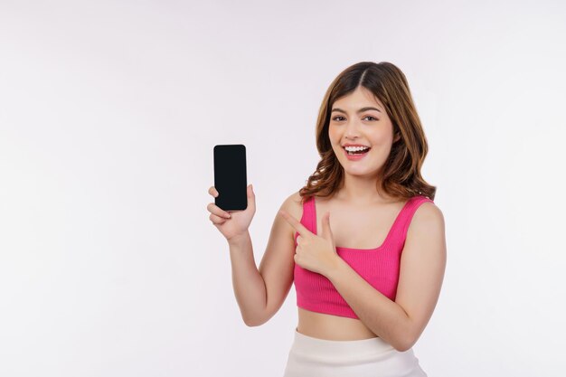 Retrato de una joven emocionada sosteniendo un teléfono móvil simulado y señalando con el dedo al smartphone aislado sobre fondo blanco