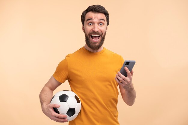 Retrato de un joven emocionado mirando su teléfono inteligente y sosteniendo una pelota