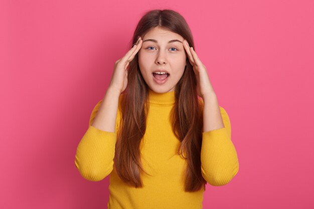 Retrato de una joven emocional conmocionada que abre ampliamente la boca y los ojos, pone las manos en las sienes, tiene dolor de cabeza, está estresada, siente dolor, tiene problemas de salud. Concepto de personas y estrés.