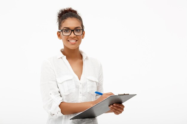 Retrato de joven empresaria africana exitosa en gafas sonriendo, escribiendo en la carpeta, mirando a la cámara en blanco