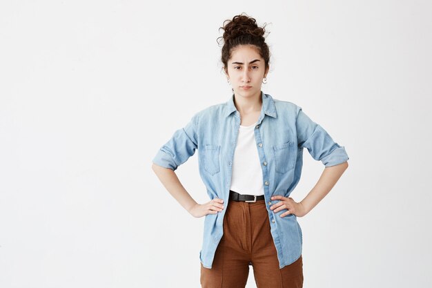 Retrato de joven empresaria con cabello oscuro en moño con ropa casual, mirando con molestia, de pie con las manos en las caderas, enojado con sus empleados que muestran resultados de trabajo insatisfactorios