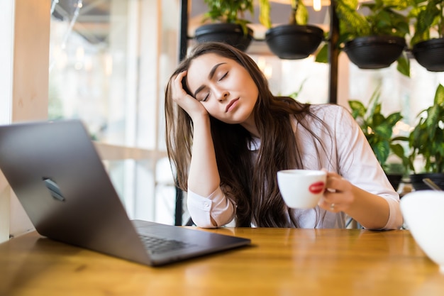 Retrato de una joven empresaria cansada sentada en la mesa con el ordenador portátil mientras sostiene una taza de café y duerme en un café