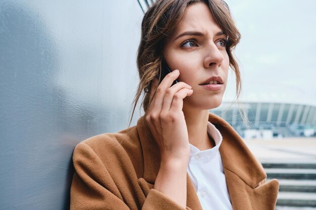 Retrato de una joven empresaria pensativa con abrigo hablando por celular al aire libre