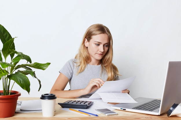 Retrato de joven empresaria trabajando con la computadora portátil y la calculadora, mirando atentamente los documentos, calculando las facturas de la empresa, haciendo informes financieros. Concepto de personas, carrera y negocios