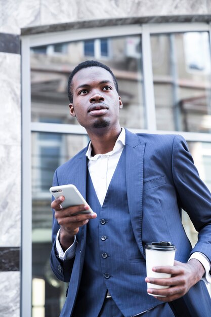 Retrato de un joven empresario africano en traje azul que sostiene una taza de café para llevar usando un teléfono móvil