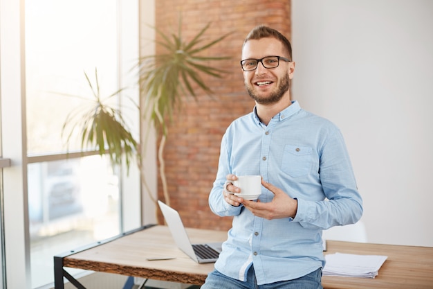Foto gratuita retrato de joven empresario barbudo con gafas y ropa casual, de pie en la brillante oficina de coworking