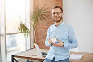 Foto gratuita retrato de joven empresario barbudo con gafas y ropa casual, de pie en la brillante oficina de coworking