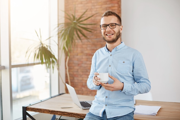 Retrato de joven empresario barbudo con gafas y ropa casual, de pie en la brillante oficina de coworking