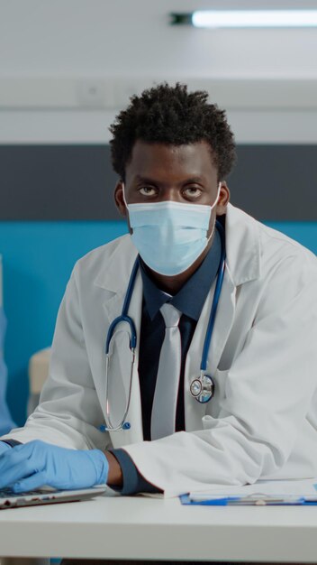 Retrato de joven especialista con ordenador portátil sentado en el escritorio con guantes y mascarilla en el consultorio médico. Doctor escribiendo en el dispositivo y enfermera con la computadora en segundo plano durante la pandemia