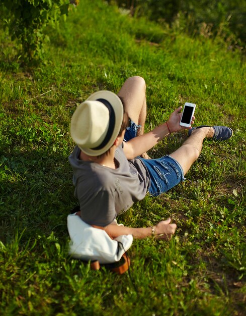 Retrato de joven con estilo moderno y atractivo hombre en ropa casual con sombrero en gafas sentado en el parque en la hierba verde mirando el teléfono
