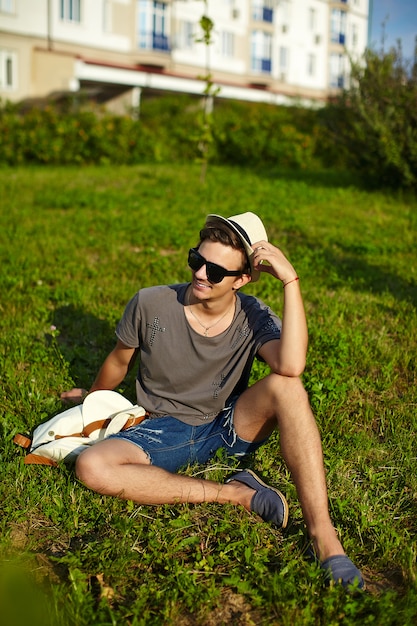 Retrato de joven con estilo moderno y atractivo hombre en ropa casual con sombrero en gafas sentado en el parque en la hierba verde