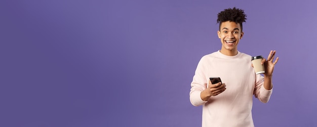 Foto gratuita retrato de un joven estudiante alegre con una taza de café y un teléfono móvil saludando con la mano para decir hola sm