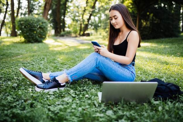 Retrato de joven estudiante asiática hablando por teléfono móvil, mirando la pantalla del portátil, al aire libre